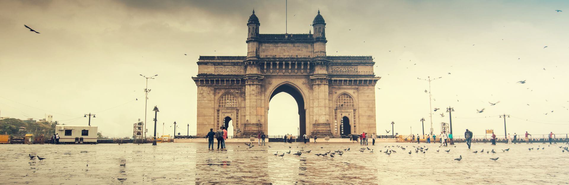 Porta dell'India a Mumbai sotto un cielo nuvoloso, con persone e piccioni sulla piazza bagnata